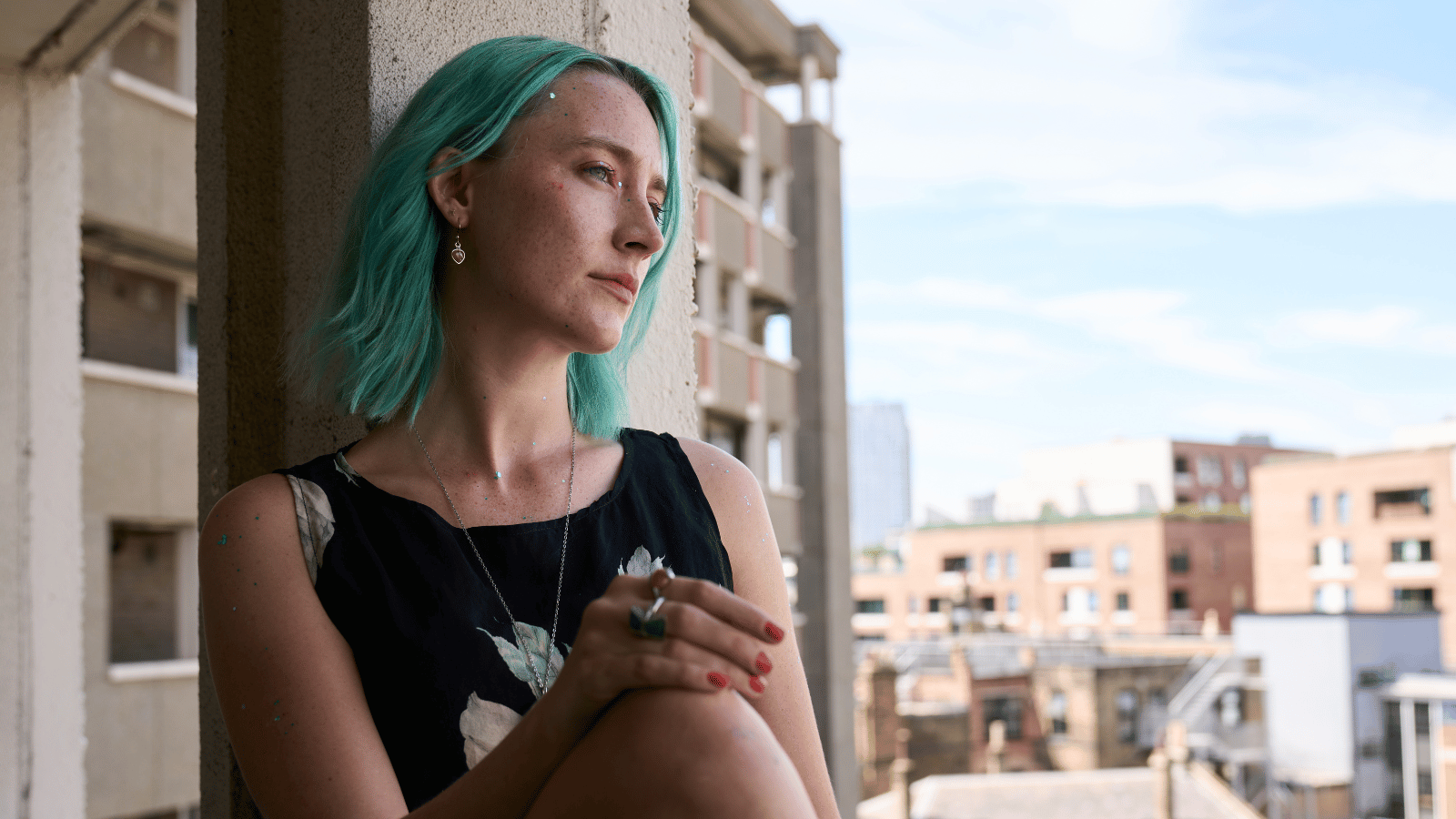 The Outrun first look still. Saoirse Ronan as Rona, a blue haired girl sitting on a window sill looking out on a building estate.