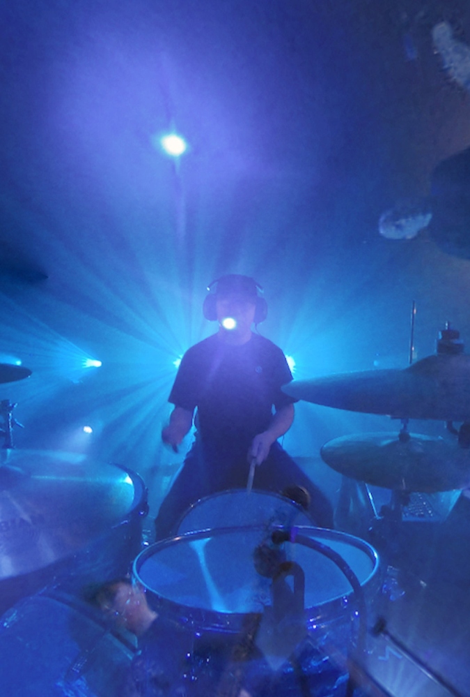 A drummer sits at a drum kit, surrounded by blue lighting on stage