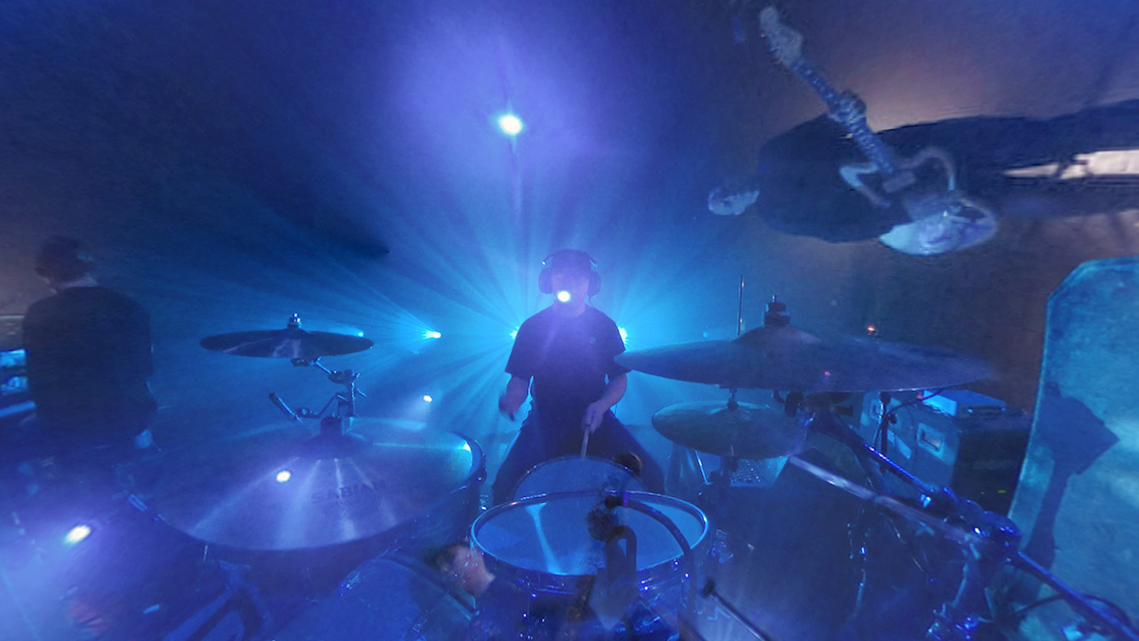 Still from Mogwai: IF THE STARS HAD A SOUND. Drummer Martin Bulloch with his kit during a performance. He is lit from behind and surround by vibrant blue light.